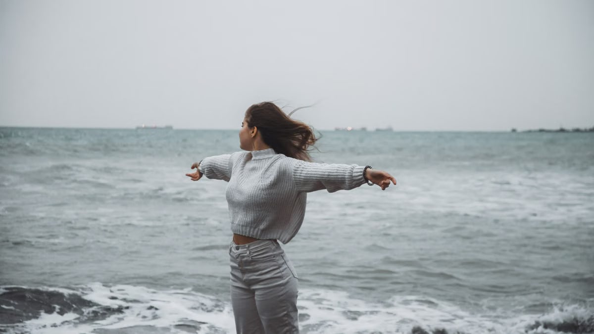 A woman stands with arms spread by the sea, embracing the freedom of a windy, overcast day.